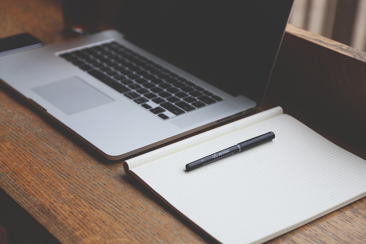 An open laptop with a blank screen is on a desk, next to it is a notepad with a pen on top of it