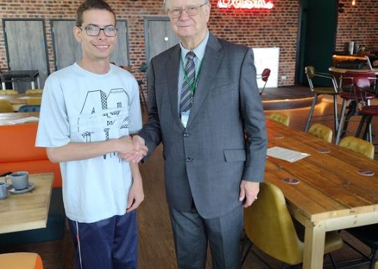 Nathaniel Yates, a white young man, dressed in purple tracksuit boots with a light blue t shirt, and black trainers. Shakes the hand of Stephen Brookes, an older white man with grey hair in thin square rimmed glasses. Stephen is wearing a dark grey suit w