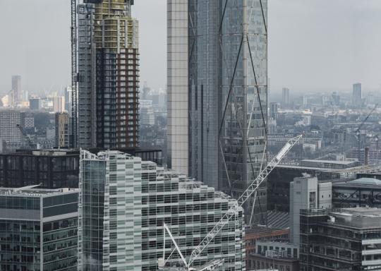 Photo of a umber of green/blue skycrapers across the London skyline. They are tall sleek buildings predominantly clad in glass. The sky is blue, and you can see a number of cranes.