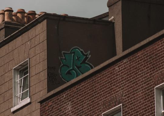Top corner of a brown brick tower-block. You can see a few white square window frames, and two brown terracotta chimney stacks. The sky is dark and grey. There is a a pink graffiti tag of the letters JB on a far corner wall at the top of one of the blocks