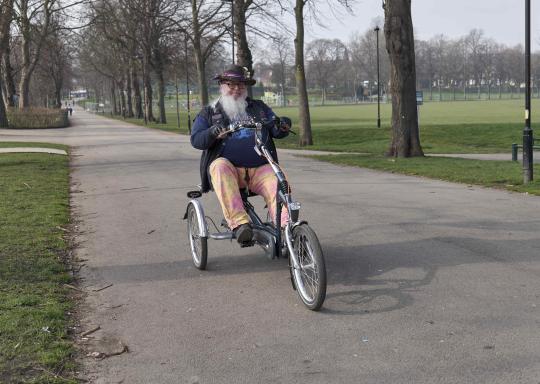 An older white man rides an adapted tricycle along a tarmac path inbetween a long line of trees in a green park. They are smiling, with a long grey beard and are wearing a blue jumper with pale yellow trousers.