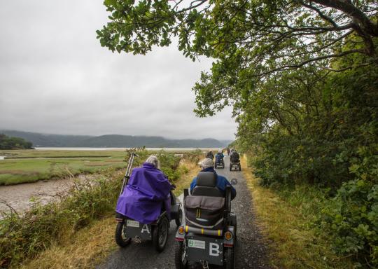 A group of older powerchair users are wheeling down a secluded rural path. They have large rain coats over the backs of their powerchairs. The path is surroduned by lush green space, including a dipped valley with a river. The sky above is grey and cloudy