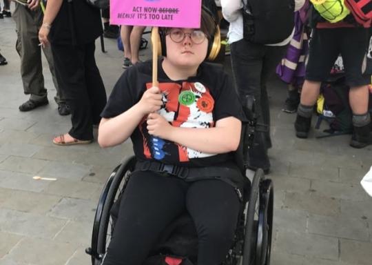 Image shows young climate change activist seated in her wheelchair. She is wearing glasses and has noise reduction headphones on. She is holding up a pink sign which says: Extinction Rebellion, act now before it is too late.