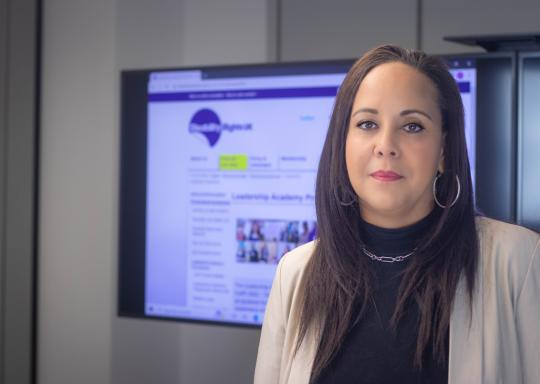 Photo of Katrina, who is waering a black polo neck and grey blazer, standing in front of a powerpoint presentation, looking into the camera
