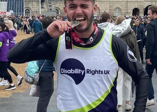 Photo of Mitch, a runner, wearing a Disability Rights UK running vest smiling and biting his marathon medal