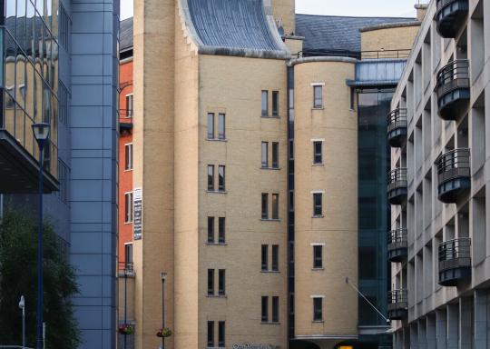 One Temple Quay in Bristol's Temple Meads are. It is a pale orange office building, with narrow cobbled street lined with grey metal benches in front of it. You can see the pale orange building in the middle of two other large office buildings.