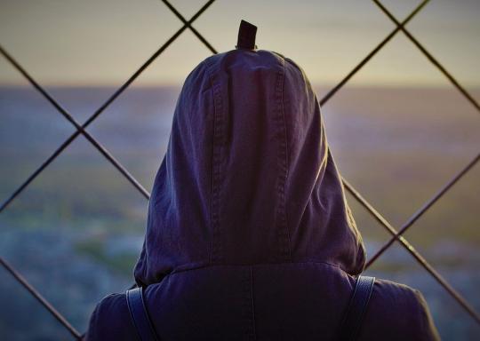 image shows the back of the head of someone wearing a hoodie. They are looking through a wire fence into the distance.