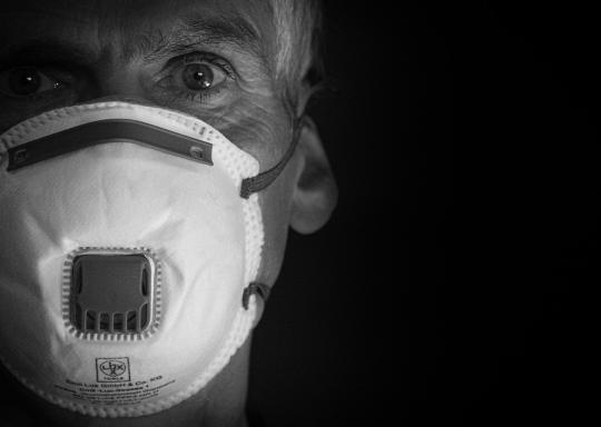image is a black and white of a close up of a mans face. He is wearing a face mask covering his nose and mouth.