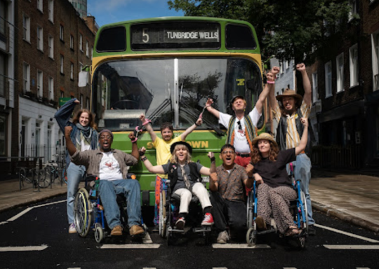 Image shows the cast as a group of eight disability campaigners, three in wheelchairs, blocking the route of a Tunbridge Wells bus in the mid-1990s.