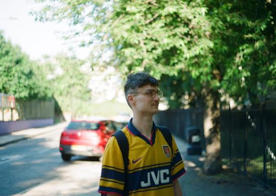 Mikey is a young white man. He is facing slightly to the side on a London street. He wears a yellow and blue striped sports top, has a short back and sides haircut and wears round thick framed glasses.