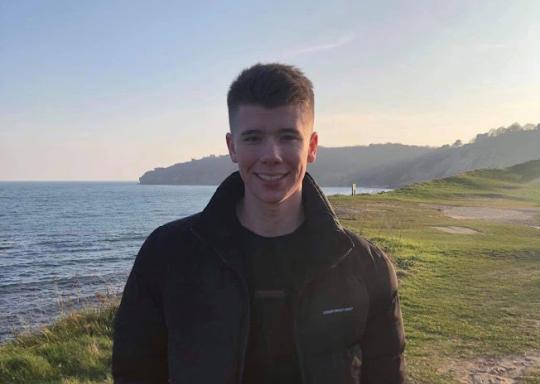 Ned Kelly is a white 19 year-old with short brown hair. He wears a brown anorak style jacket over a black t-shirt and is smiling into the camera. He is standing on grass with a body of water behind him.