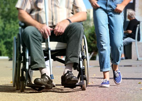 Image shows a man in a wheelchair and a carer walking alongside him. The image is cropped so only the legs of both are shown. It is a daylight scene.