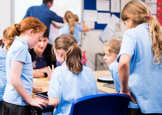 Children in a classroom standing round a table with their backs facing the camera, working on something together