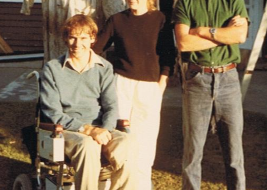 Photo of three people in a garden, one man in a wheelchair, a woman and a man standing. A washing line with clothes hanging on it can be seen behind them