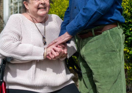 Image of a woman smiling and sitting in a mobility aid whilst she holds the hands of a man standing next to her