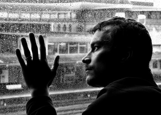 Image shows man with head and hand pressed against a rain soaked window. Image is black and white