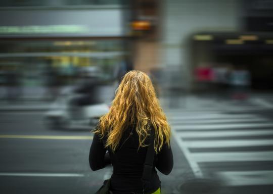 image shows the back of a girls head as traffic races by. She has blonde hair and a black jacket. The passing traffic is blurred.