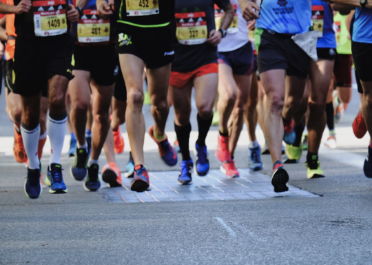 Photo of the legs and feet of people running a marathon. They are wearing running vests with race numbers on them, and trainers