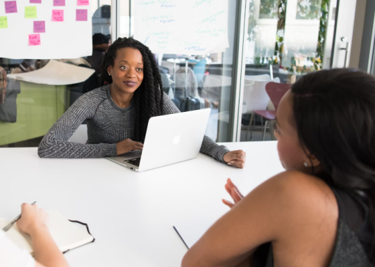 Image of three women sitting talking around a work table with post-it notes up on the wall behind them