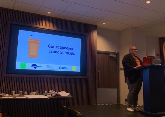 Image of Isaac talking on stage at the moving social work event next to a presentation that says 'Guest Speaker Isaac Samuels'