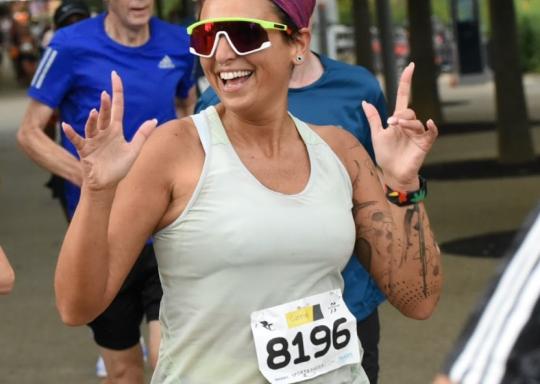 Image of Manuela in running gear, smiling and throwing her hands up in delight as she runs the course