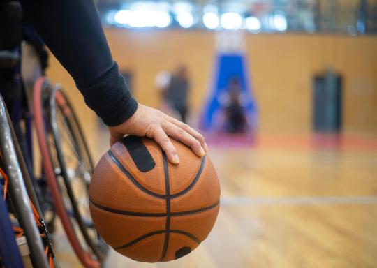 image of a basketball held in the hand of a wheelchair user in the middle of a wheelchair basketball game