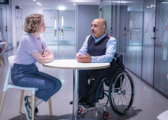 Leanne, a white woman with a short blonde bob haircut, is sat across a white table from Kamran Mallick, a south asian man who is bald and a wheelchair user. Leanne is wearing a light blue t shirt tucked into blue jeans. Kamran is wearing black trousers an