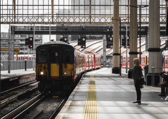 Photo of a train coming into the platform at Waterloo station. The train tracks are to the left, with a roof over the station. There is a woman standing on the platform to the right hand side