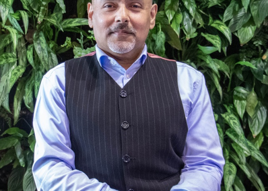 Kamran is smiling, he is sat in a wheelchair in front of green leaves