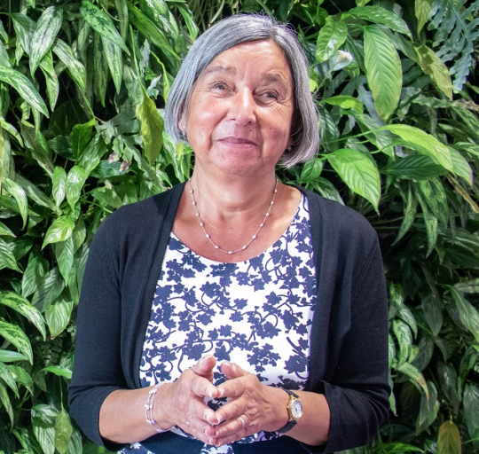 Fazilet has short grey hair and is smiling as she stands in front of green leaves