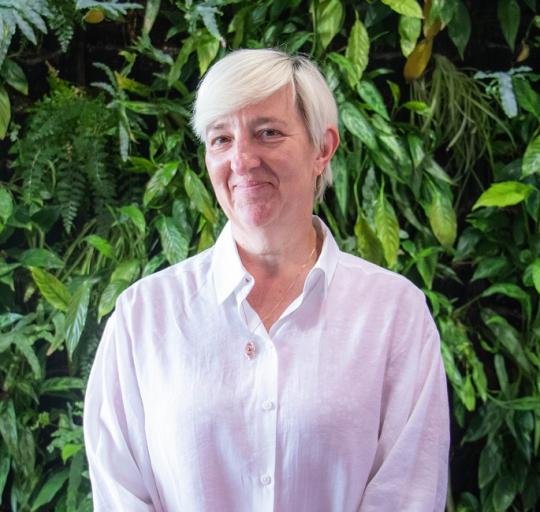 Rebecca is female, she stands in front of a green plant background and is smiling.